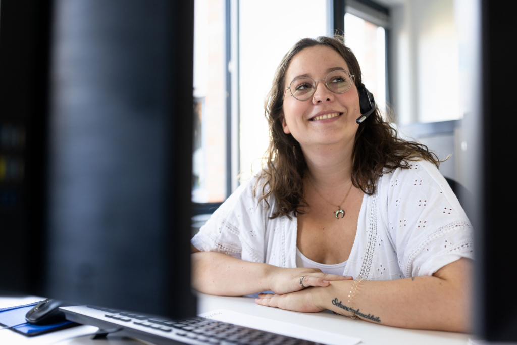 Fröhlich lächelnde Mitarbeiterin an Schreibtisch. Arbeitsproben aus dem Bereich Corporate- und Employer Branding Fotografie.